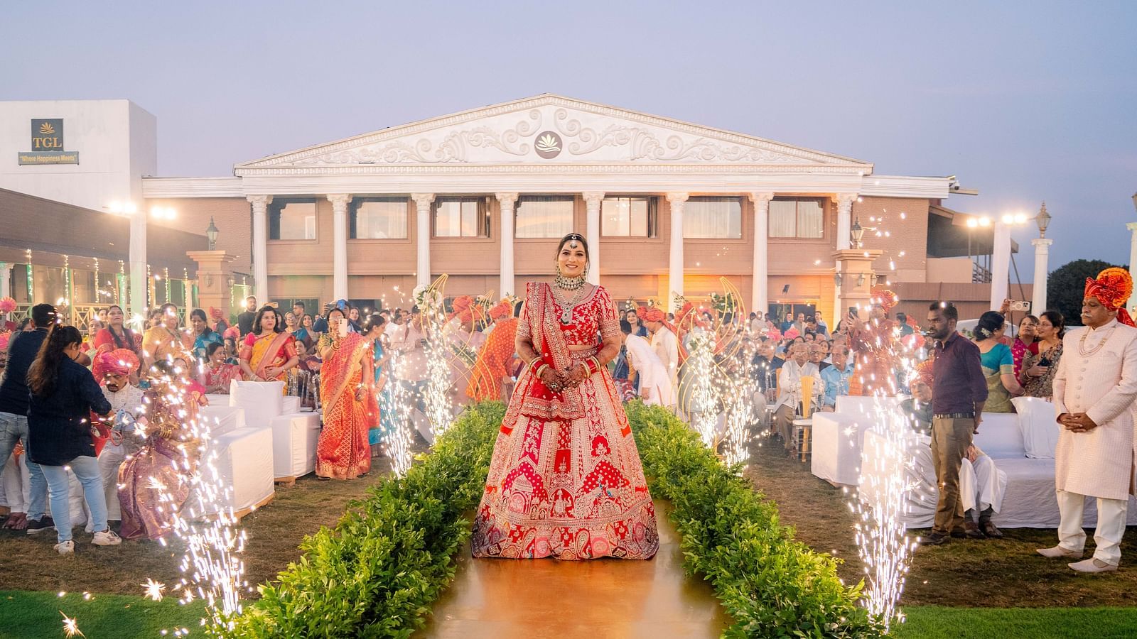A wedding procession with the bride and groom walking on a flower-lined path, accompanied by people and celebratory sparklers. A large building is in the background - The Grand Legacy Resort and Spa