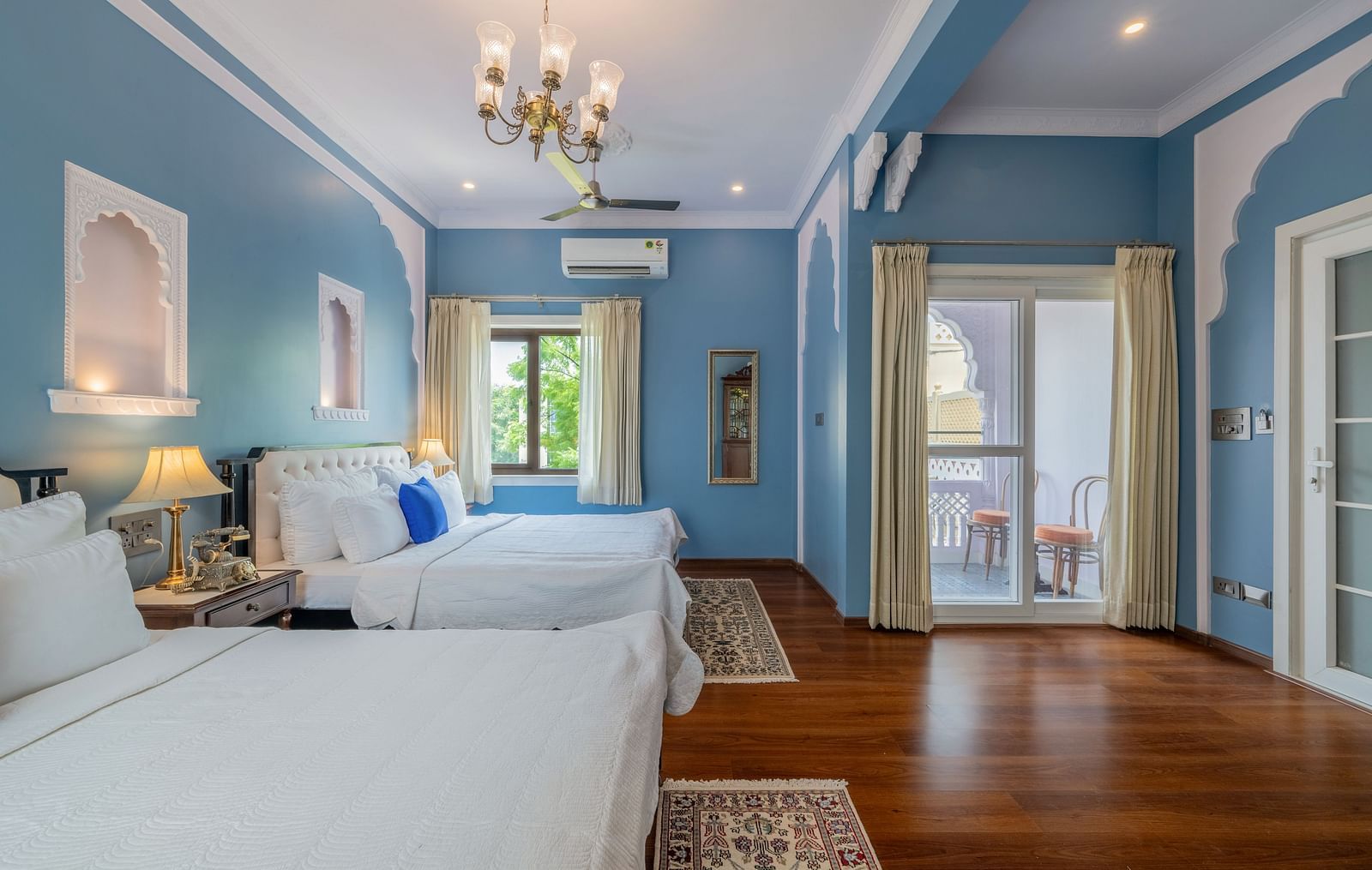 A bright and airy room at Dera Rawatsar, Jaipur, offers two comfortable beds, a balcony with seating, elegant wall details, and wooden flooring.