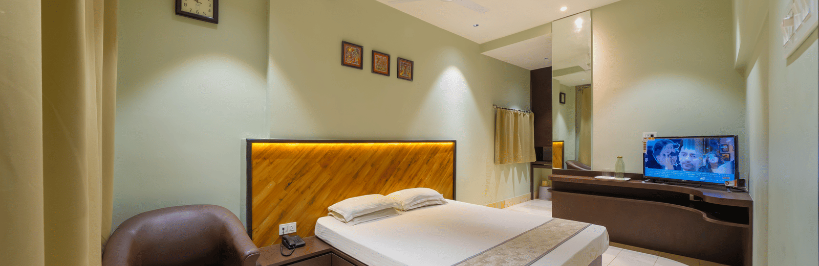 A spacious hotel bedroom featuring a double bed with a headboard, light green walls, a TV, and a leather armchair - Hotel Dolphin Puri