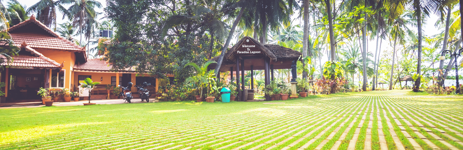 Lawn with palm trees, tiled pathways, and red-roofed building surrounded by greenery at Paradise Lagoon Resort, Udupi.