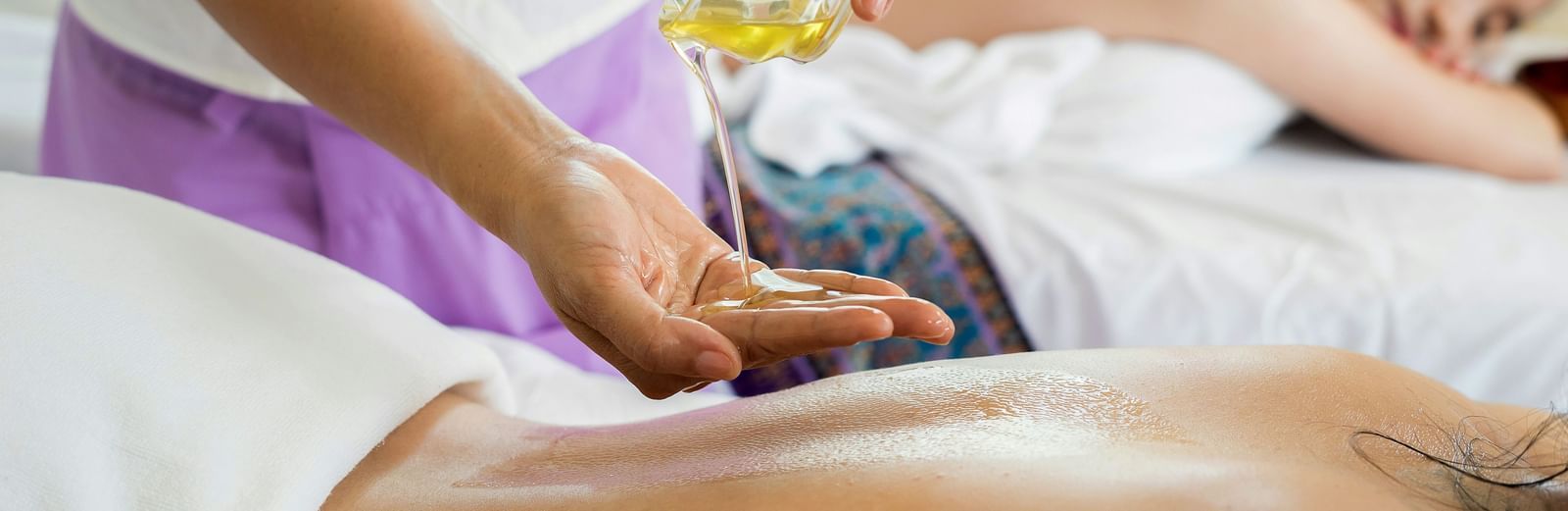 Masseuse applying oil to a person's back during a spa treatment, with another person lying on a massage table in the background.