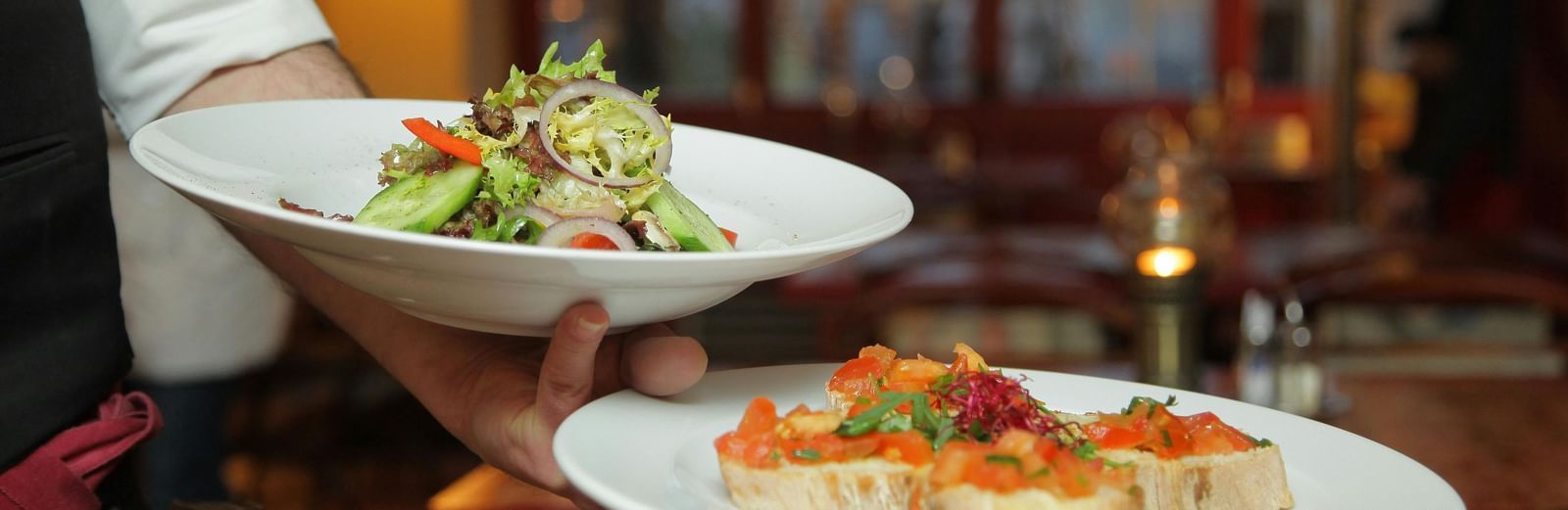 A waiter in a white shirt and black vest carefully carries a plate of salad and a plate of bruschetta in a warmly lit restaurant setting.