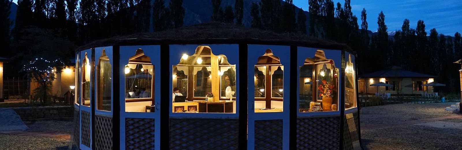 The exterior of a building with large windows, glowing warmly against a dark, tree-lined background at night at White Sands Resort, Ladakh.