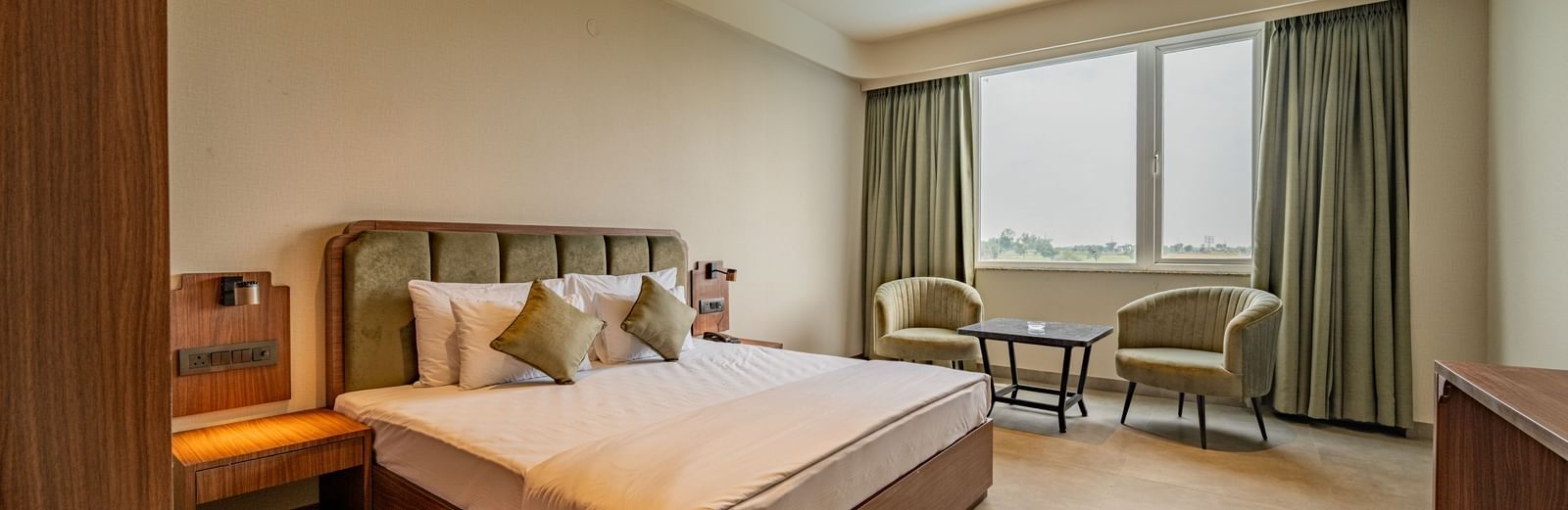Warmly lit room with a large bed, cushioned chairs beside the windows, and wooden furnishings at Hotel Highway King, Neemrana.