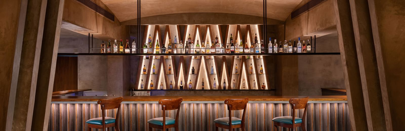 A dimly lit bar with a long counter, several bar stools, and shelves of bottles illuminated from behind, at Shilton International, Kochi.