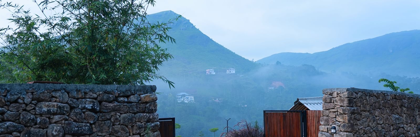 A stone wall entrance with an open wooden gate at Vaasta Vagamon, revealing a winding road and distant mist-shrouded hills.