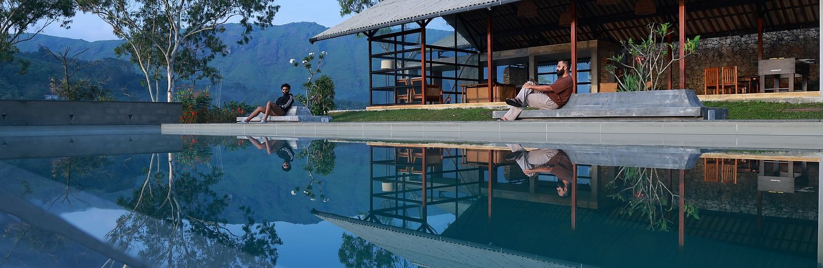 Two people relax by an infinity pool at Vaasta Vagamon, overlooking a serene landscape with rolling hills and trees.