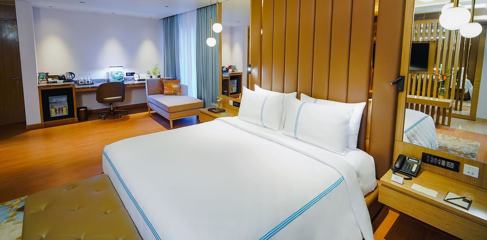 An image of a large bed under hanging lamps and a night stand on the side in our A beautiful image of a hotel room in Madurai with a large bed, a couch, and a wall-mounted television