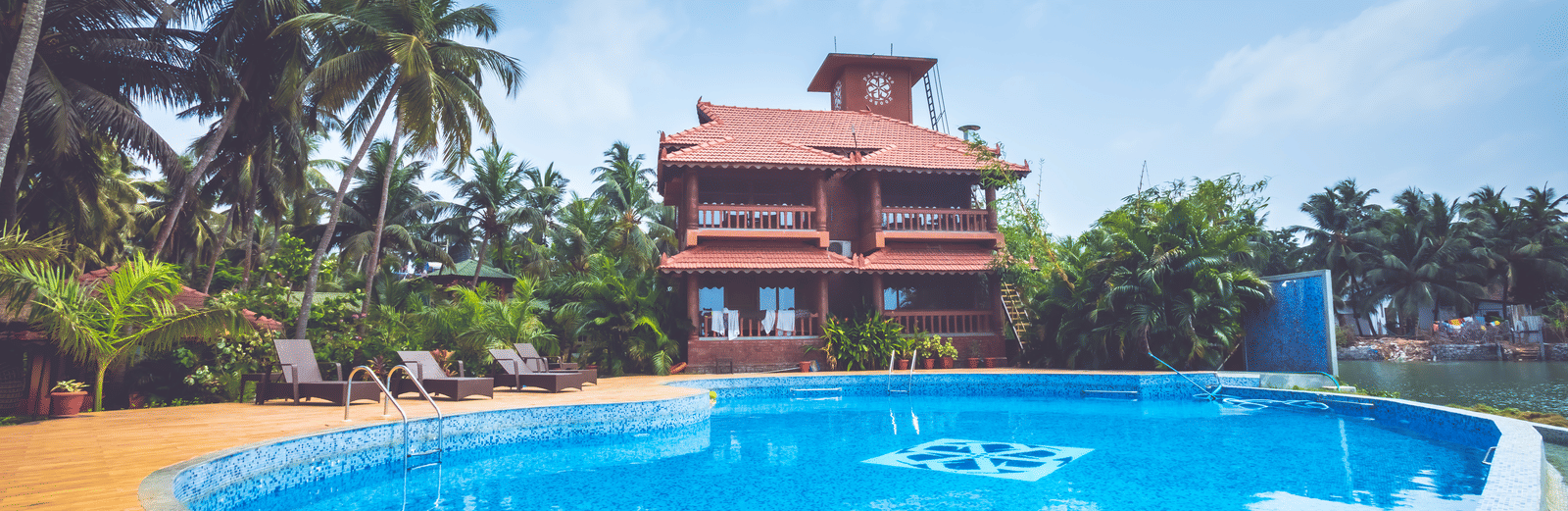 Swimming pool surrounded by palm trees, with a red-roofed house in the background at Paradise Lagoon Resort, Udupi.