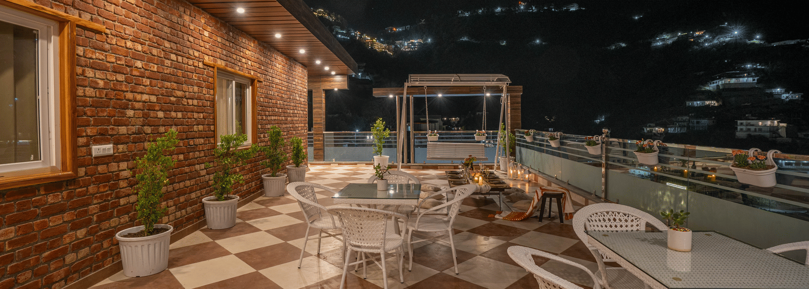 An outdoor terrace with checkered flooring, tables and chairs, potted plants, and hillside night views at Perfectstayz Value Paradise, Mussoorie.