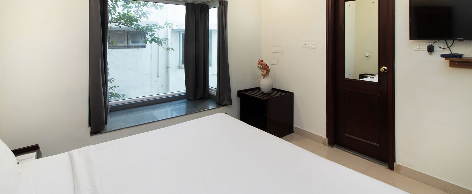 A room at Sanctum Suites Indiranagar, Bangalore showing a double bed with white linens, wooden furniture, a television, and a glass window.
