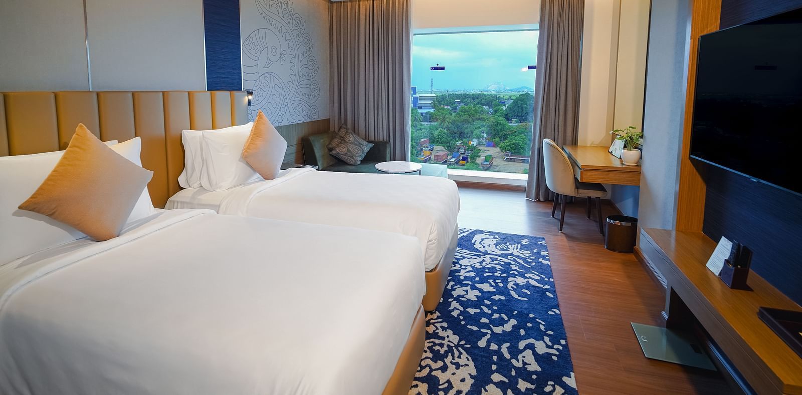 An image of a hotel room in Madurai with twin beds, a wall-mounted television, a work desk and a view of the hills