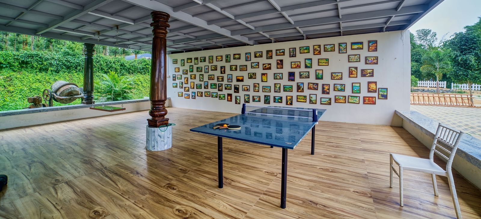 A large, covered outdoor common area with a dark tiled floor and numerous framed artworks or photos adorning a wall, with a table and chairs set up. - Vintage Trails by Ilara