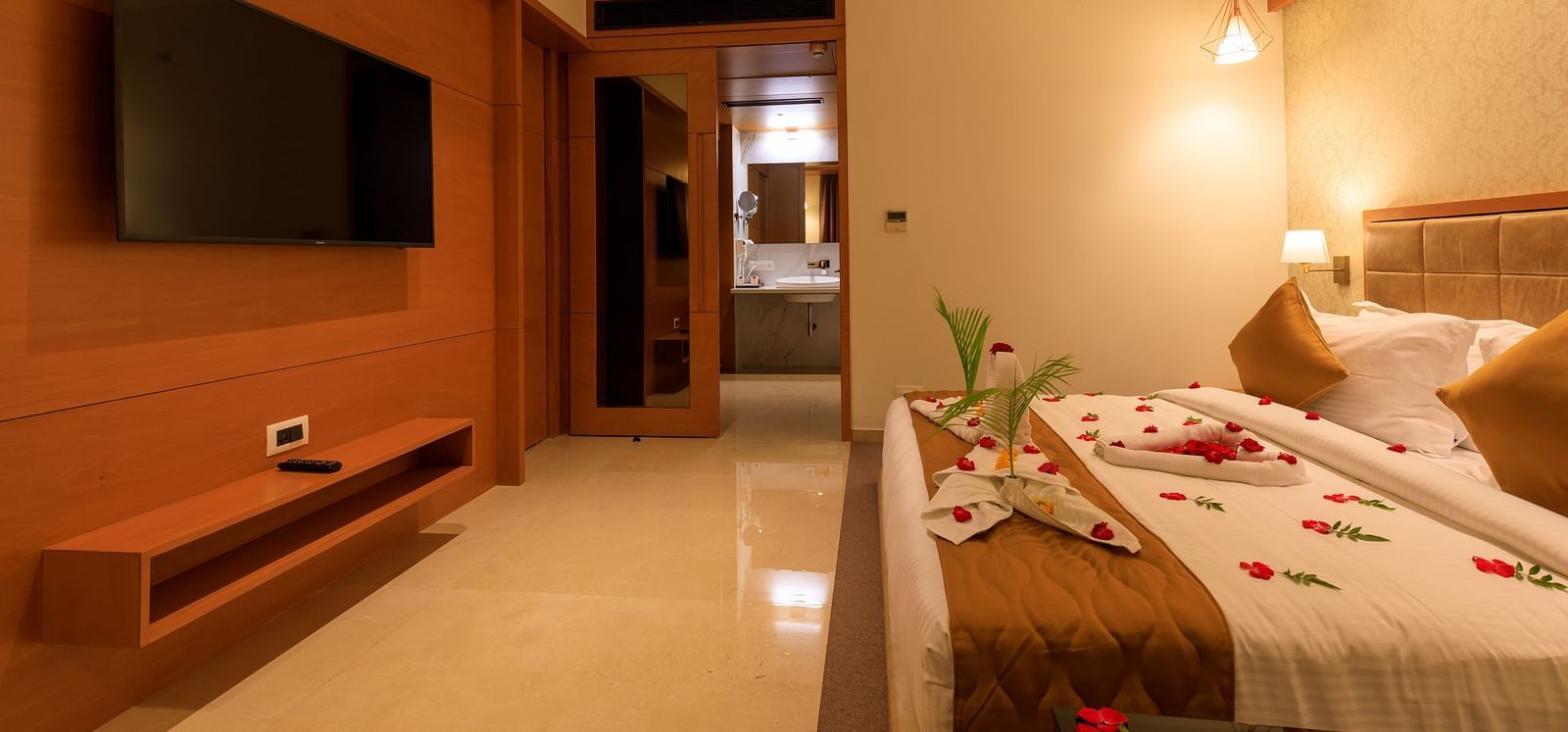 A warmly lit hotel room at Lords Eco Inn Apple Gate features a decorated double bed, a wall-mounted TV, and a glimpse into the bathroom.