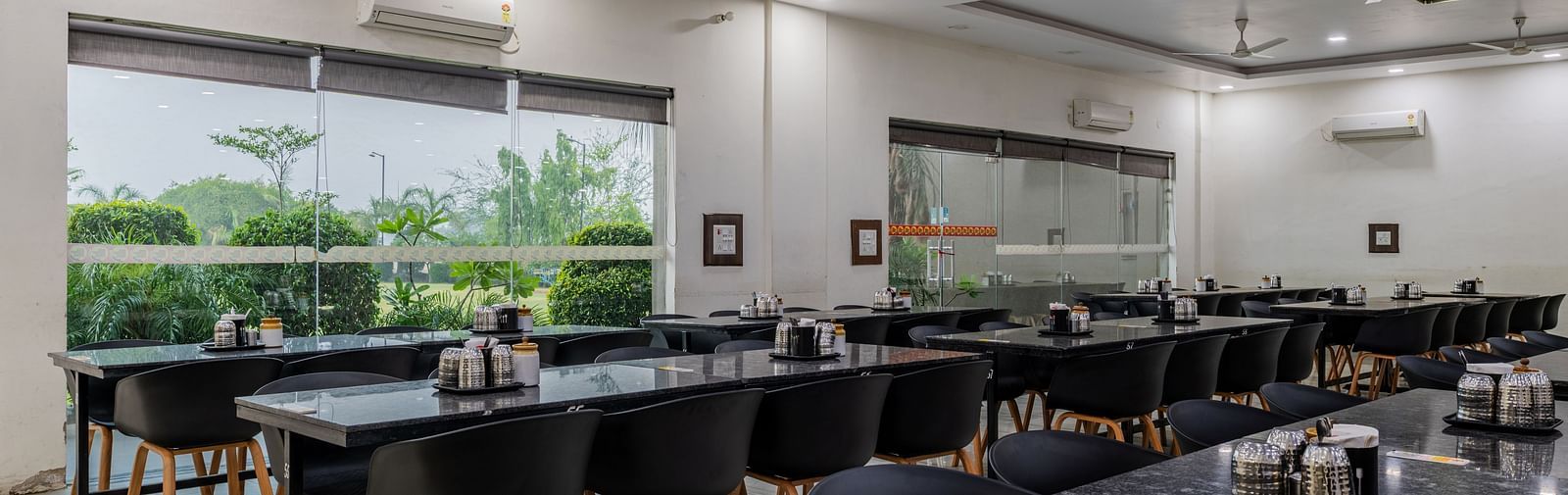 Air-conditioned restaurant of Hotel Highway King, Behror, is furnished with parallel aligned dining tables, water cups and glass windows that offer views of the trees outside at Hotel Highway King, Behror.