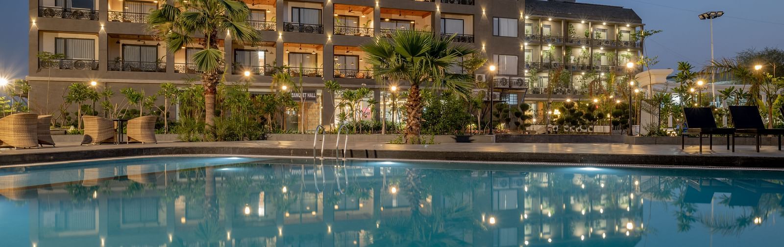 A multi-storey hotel building with colourful window accents overlooks a large, inviting swimming pool with lounge chairs at dusk.