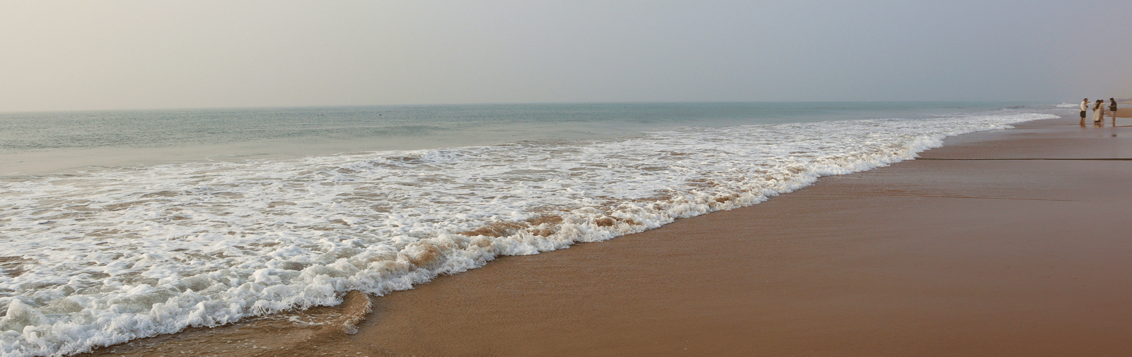 White ocean waves are breaking onto a flat, wide, sandy beach under a slightly hazy sky.