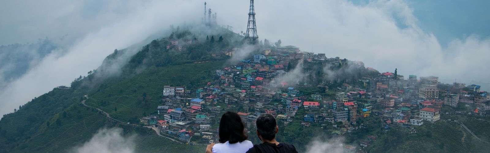 Romantic couple sitting on a hilltop overlooking Darjeeling town covered in clouds and mist, with panoramic Himalayan mountain views — best tourist attraction in West Bengal.