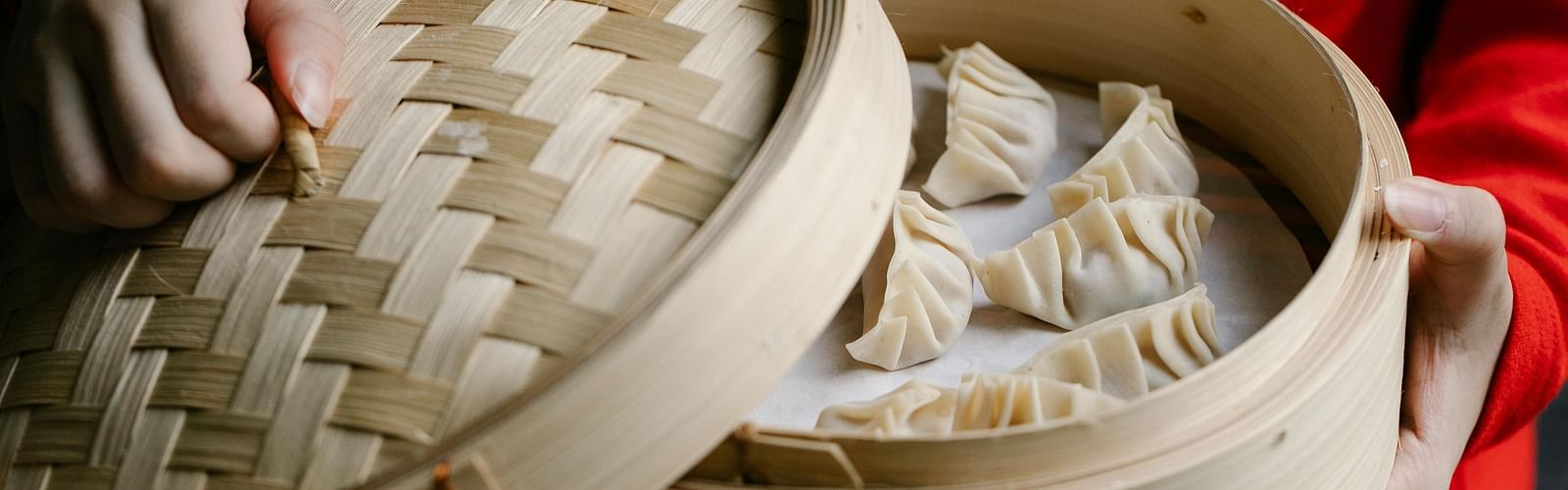 A person holding a bamboo steamer filled with dumplings, with the lid partially open