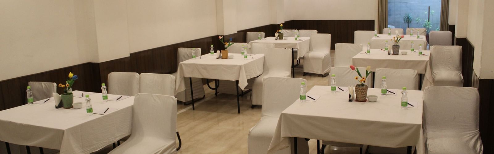 An image of a banquet hall at Inde Hotels Vista Woods featuring several sets of chairs and table where water bottles and flower pots are placed 