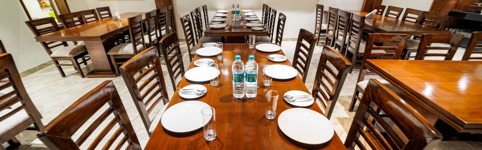 Dining area at Inde Reva Resorts Rishikesh with a long wooden table, cushioned chairs, framed artwork, and a neatly arranged dining setup.