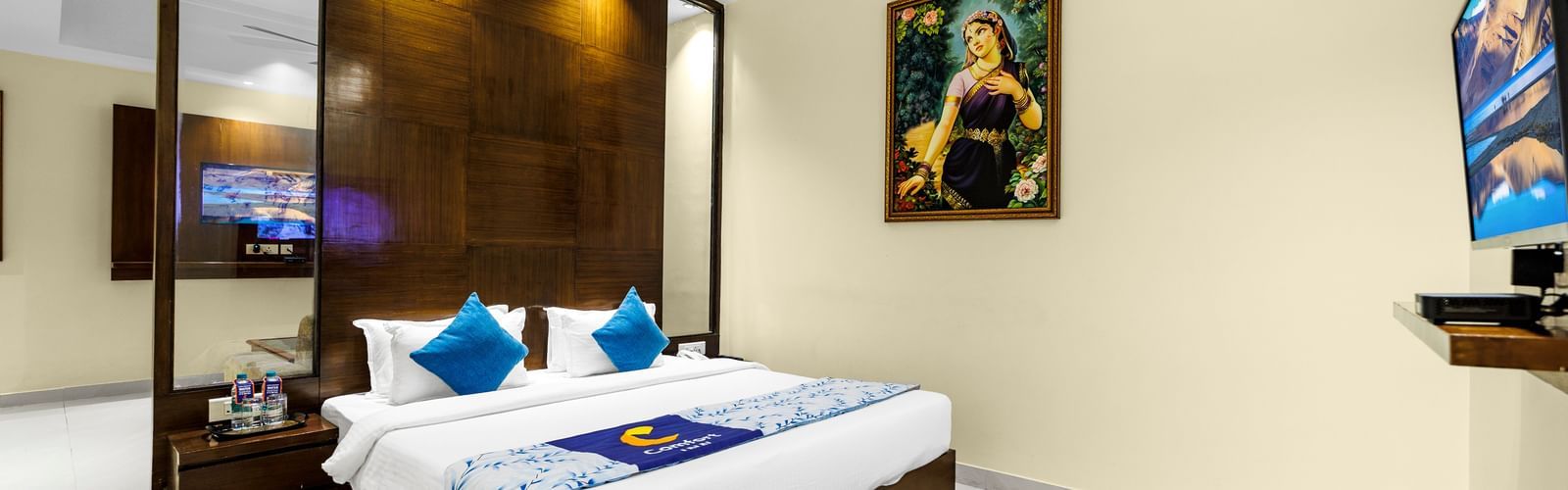 Bedroom at Inde Reva Resorts Rishhikesh with double bed, blue cushions, ceiling fan, TV, and framed artwork on the wall.
