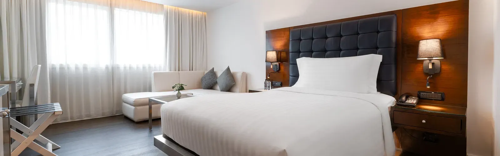 Modern hotel bedroom with tufted navy headboard, platform bed, white seating area, walnut accents, and floor-to-ceiling curtains at Night Hotel Bangkok.