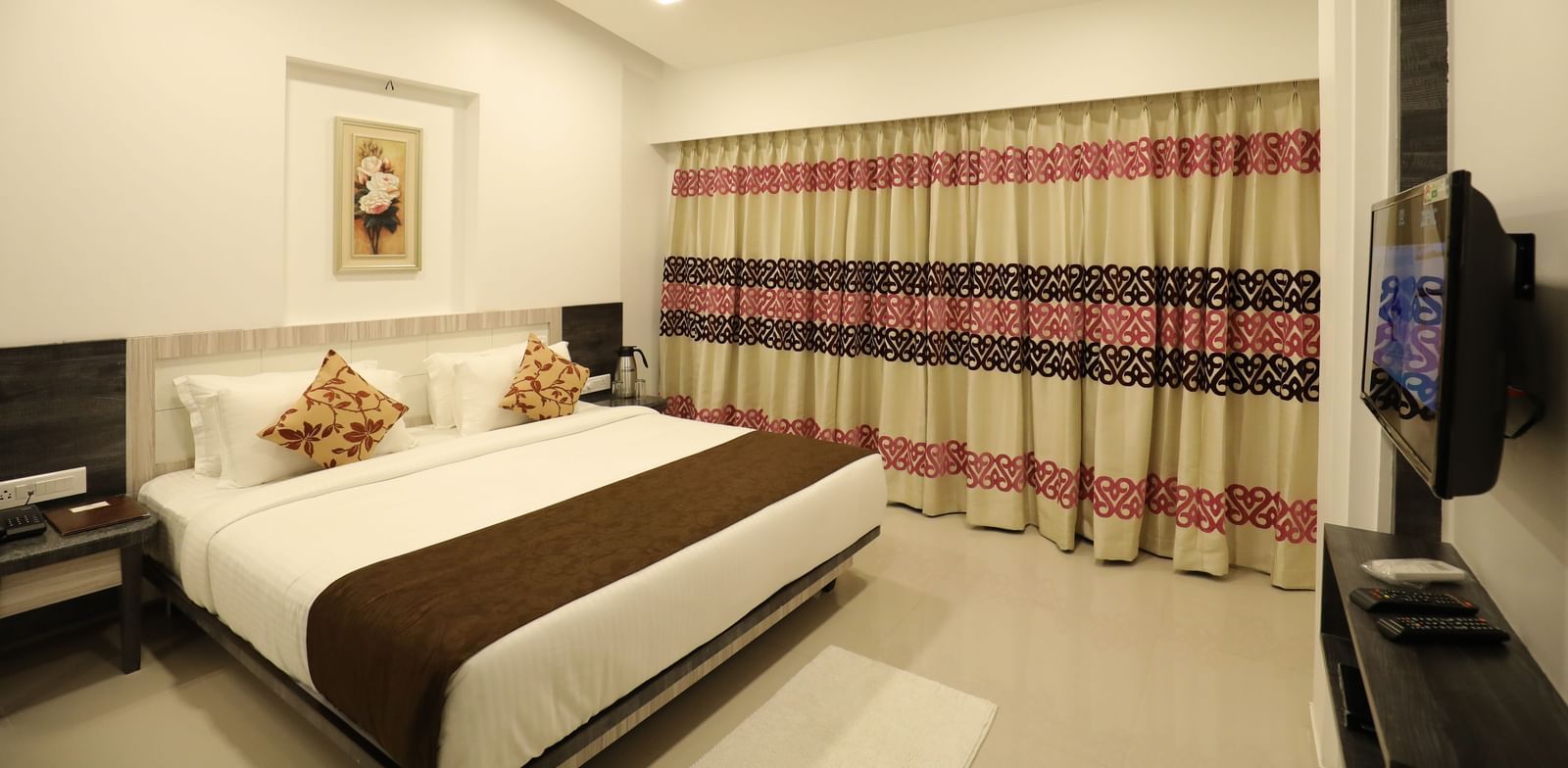 A wide view of a bedroom at Imperial Grande showing a bed with white and brown bedding, a television, and striped curtains.