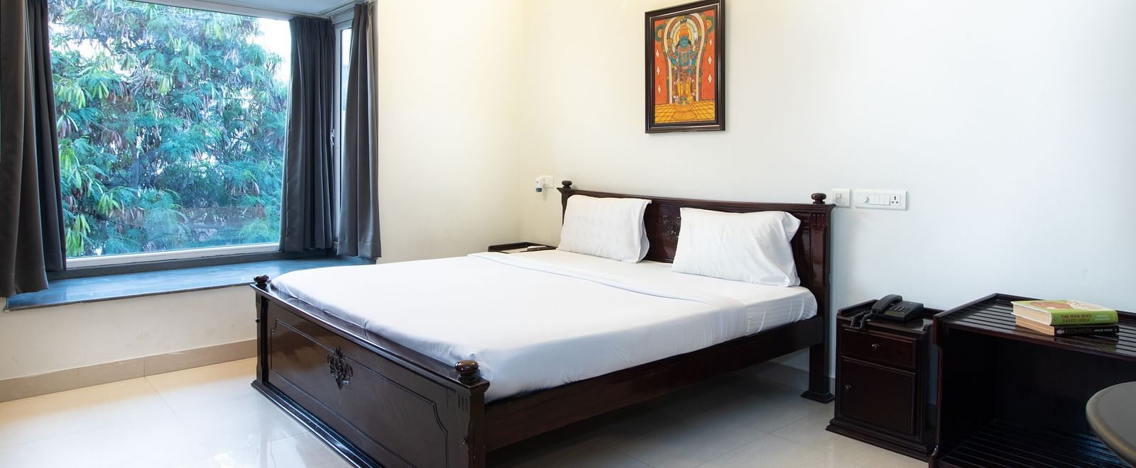 A spacious room featuring a double bed with white linens, bed side tables, and a glass window overlooking greenery at Sanctum Suites Indiranagar, Bangalore.