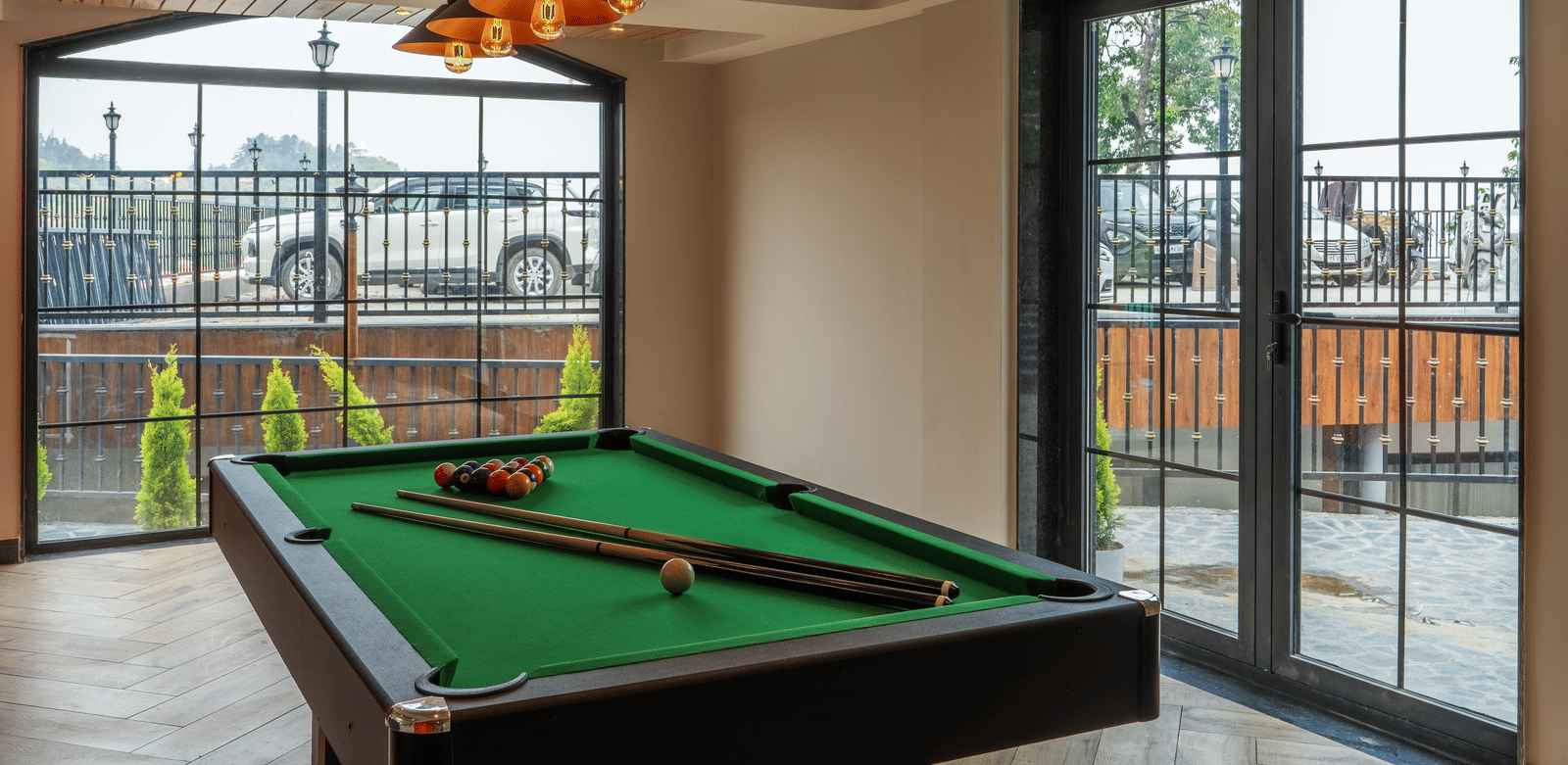 A green billiard table with cues, inside a room with large glass windows and hanging lights at Perfectstayz Luxe Mall Road.