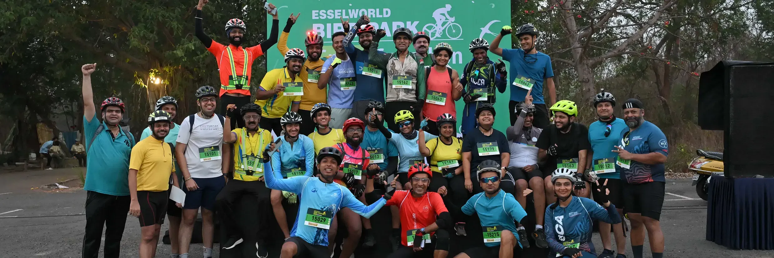 Cyclists pose together after a successful event, celebrating in front of a green Cyclothon banner at an outdoor venue surrounded by trees.