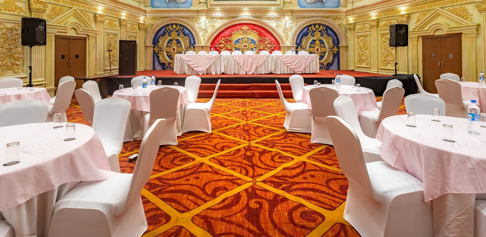 Elegantly decorated banquet hall at Diamond Palace Lords Plaza set with round tables and chairs facing a stage.