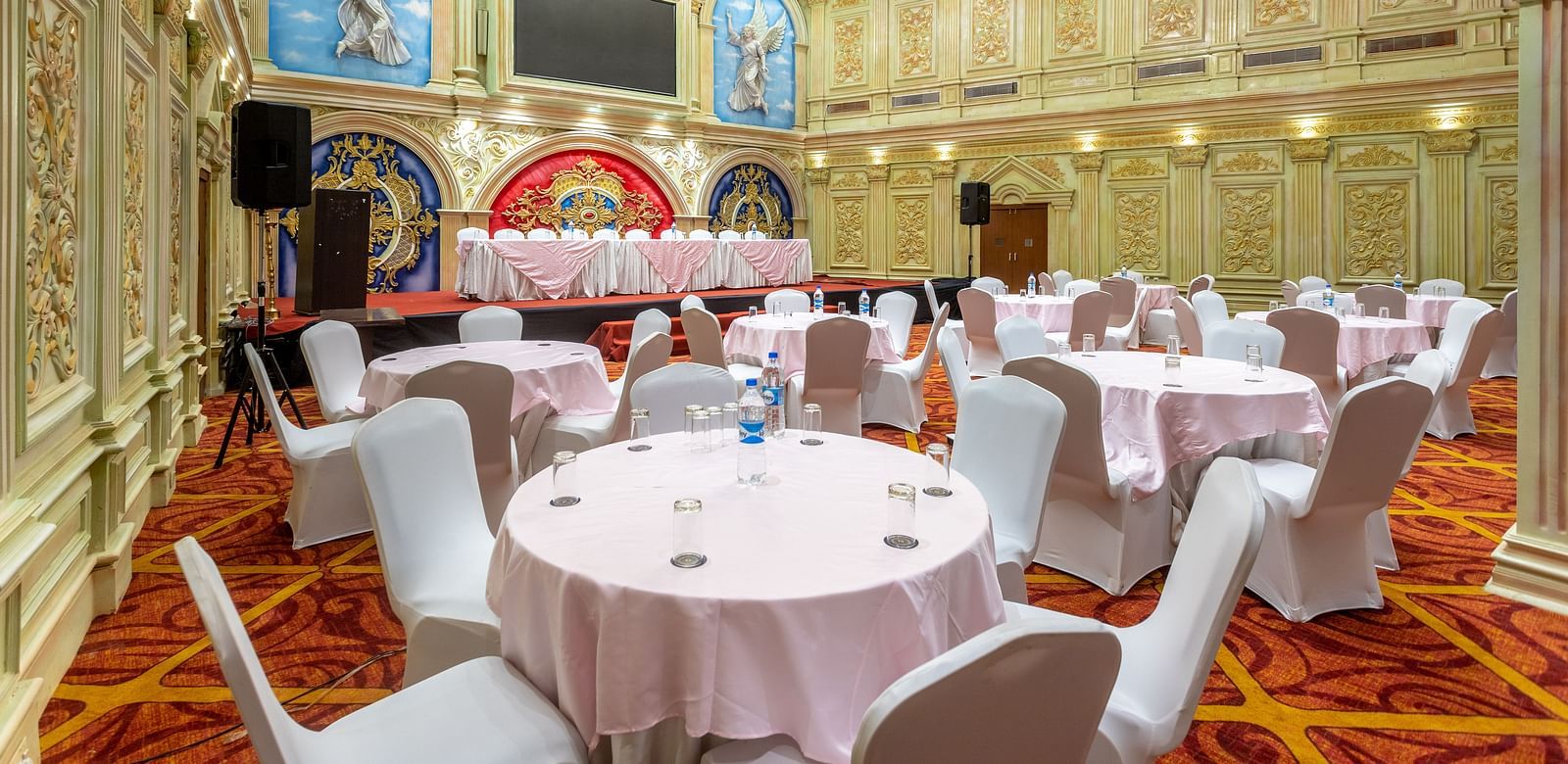 Ornate banquet hall at Diamond Palace Lords Plaza with round tables, chairs, chandeliers, and decorative walls.