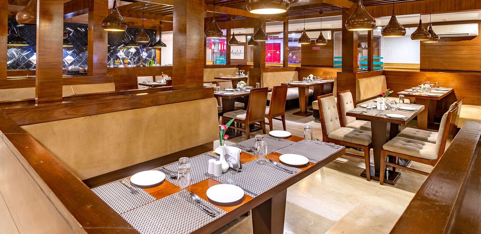 Warmly lit restaurant at Diamond Palace Lords Plaza with wooden beams, set tables, and booth seating.