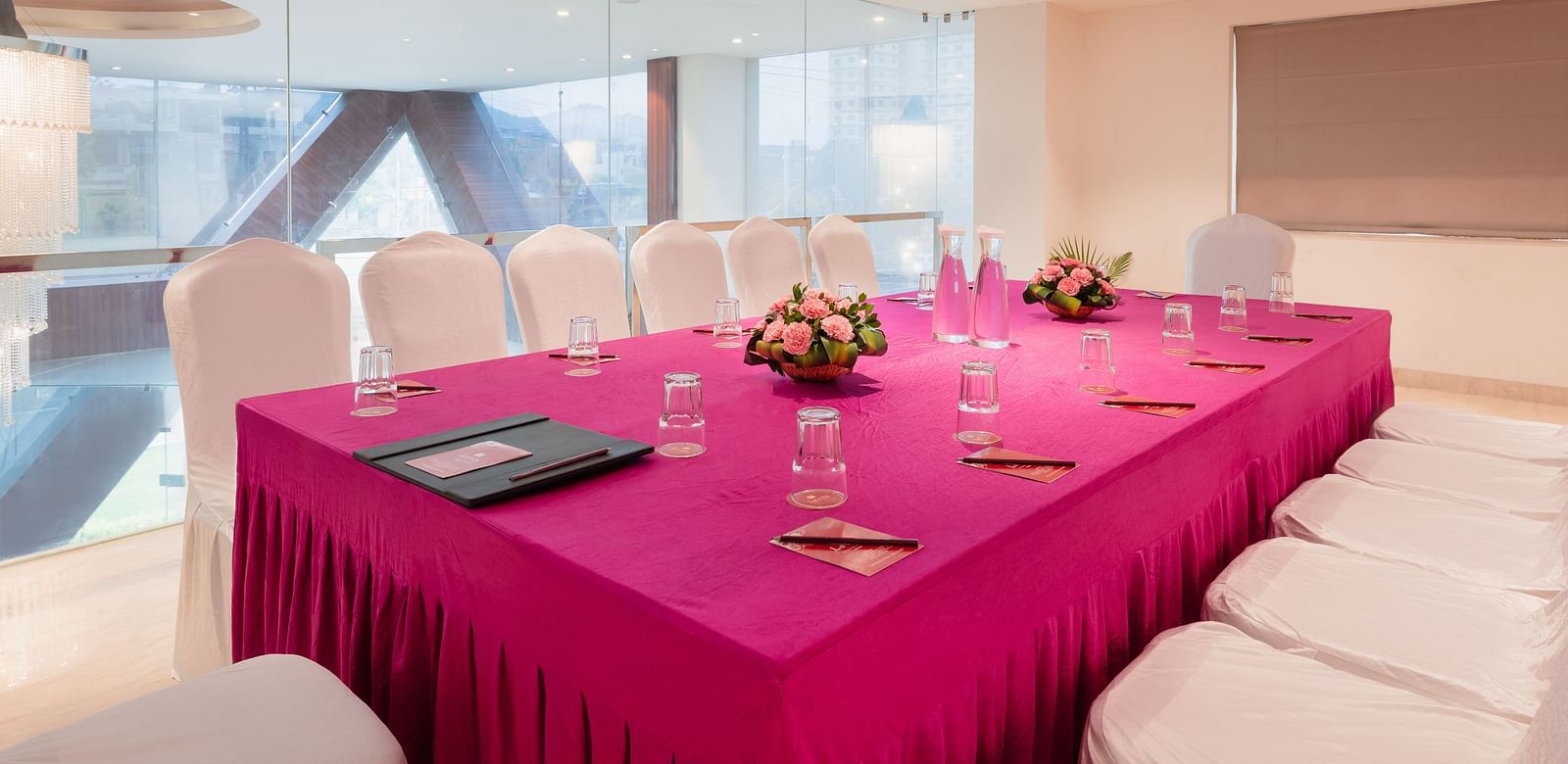 A meeting room at Lords Inn Udaipur is set with a long table covered in a vibrant cloth, surrounded by white chairs and adorned with floral centerpieces and water glasses.