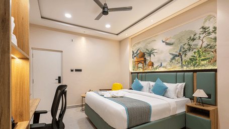 A bedroom setup displaying a king size bed with blue pillows, a desk area with a chair, and a wall mural at Monday Hotels & Suites Madhapur Hyderabad.