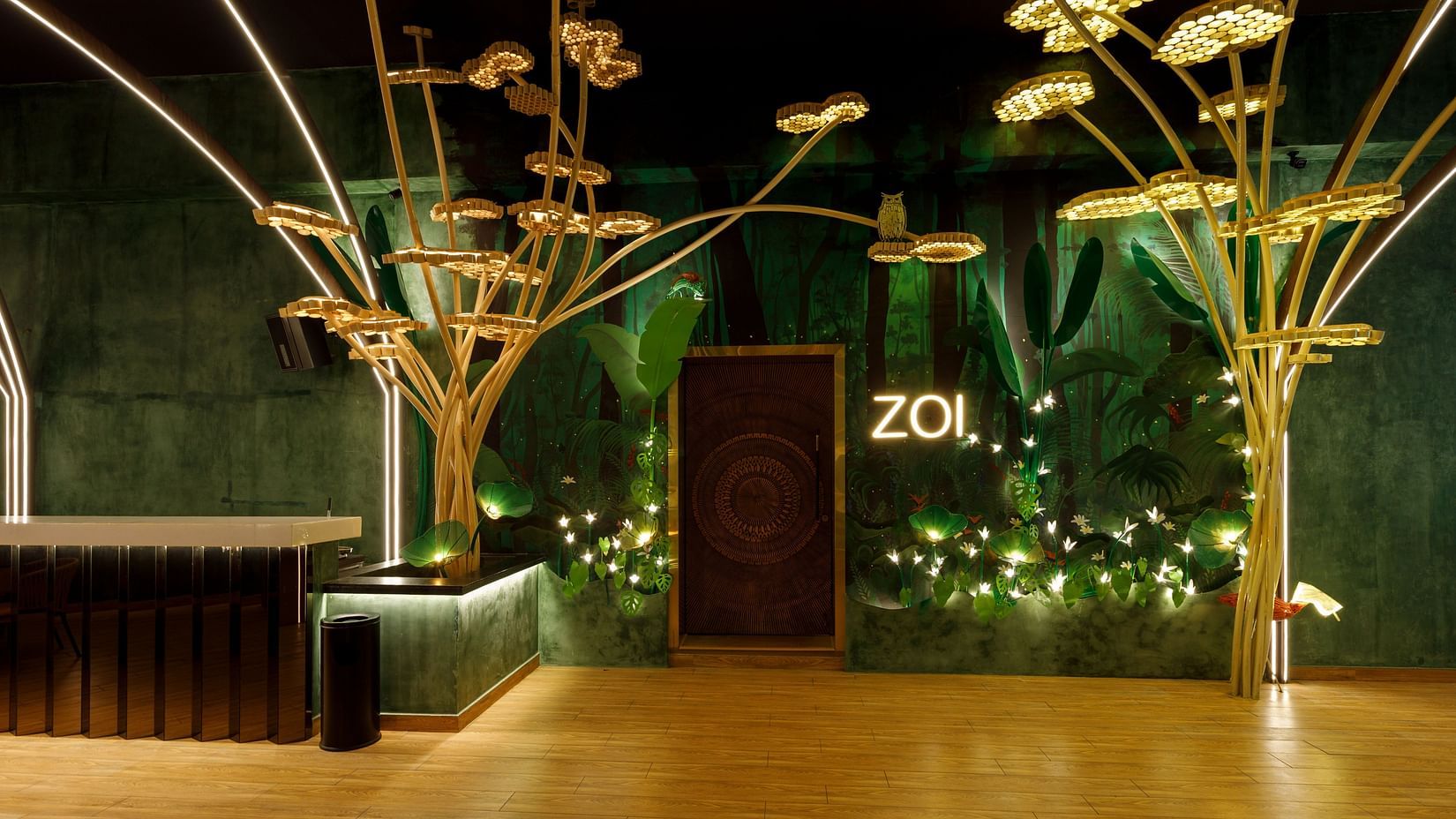 The ZOI Bar & Kitchen stylish lobby with golden decorative tree sculptures, walls, a wooden floor, a reception desk on the left, and a door with ZOL illuminated above it