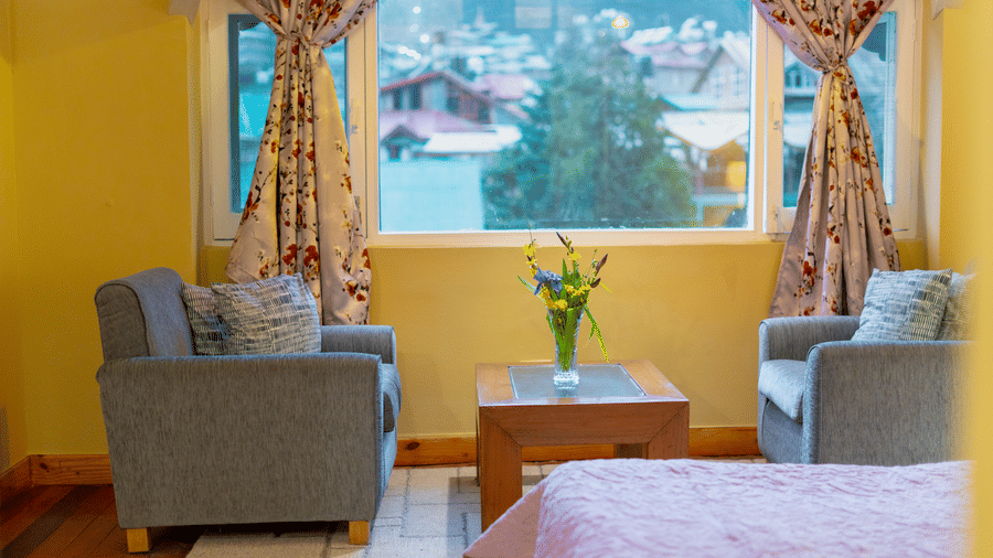 A charming sitting area with two grey armchairs and a coffee table positioned by a window with mountain views - Johnson Lodge & Spa, Manali