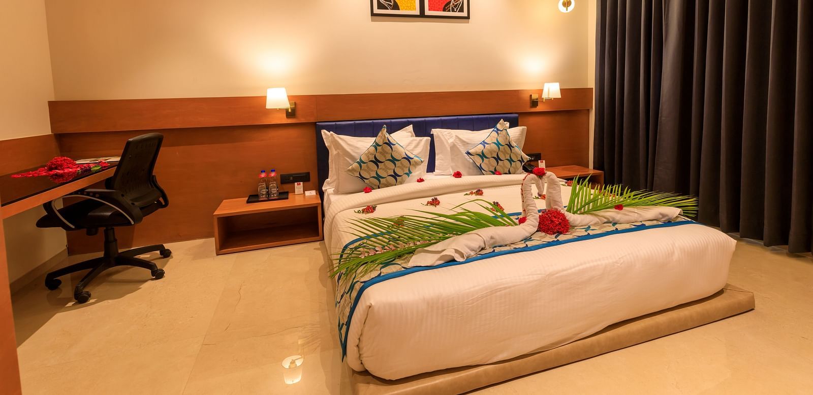 A warmly lit hotel room at Lords Eco Inn Apple Gate features a decorated double bed, bedside tables, a desk with a chair, and dark curtains.