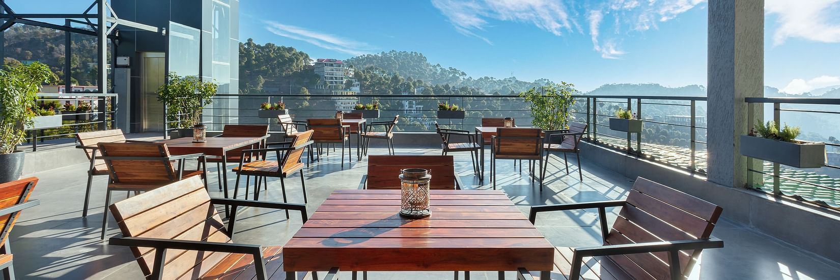 Outdoor dining area at Suryavilas Luxury Resort and Spa, featuring wooden tables and chairs on a spacious terrace with panoramic views of the Himalayan hills under a bright blue sky.
