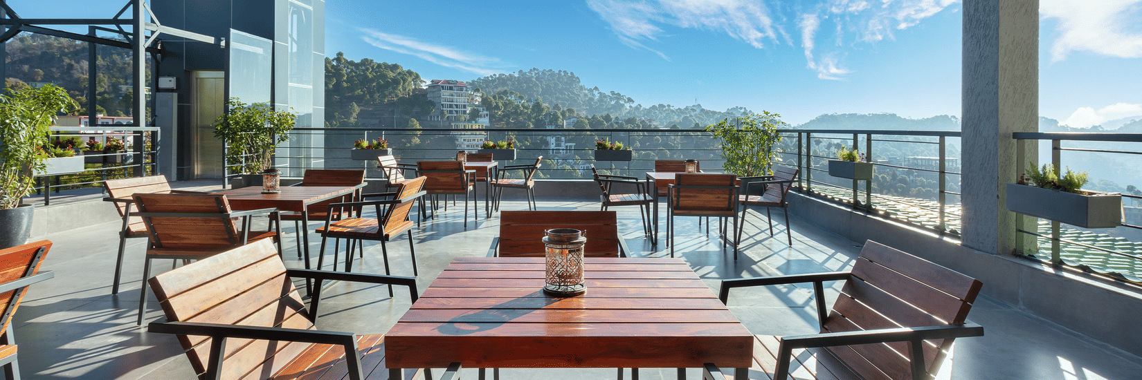 Outdoor dining area at Suryavilas Luxury Resort and Spa, featuring wooden tables and chairs on a spacious terrace with panoramic views of the Himalayan hills under a bright blue sky.