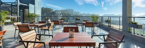Outdoor dining area at Suryavilas Luxury Resort and Spa, featuring wooden tables and chairs on a spacious terrace with panoramic views of the Himalayan hills under a bright blue sky.
