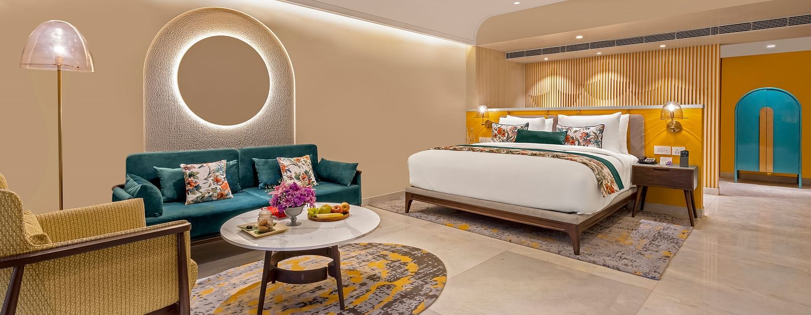 An image of a room featuring a king-size bed, a comfortable sofa, a coffee table, and a television. The room is decorated with a circular mirror, a ceiling fan, and stylish lighting fixtures. 