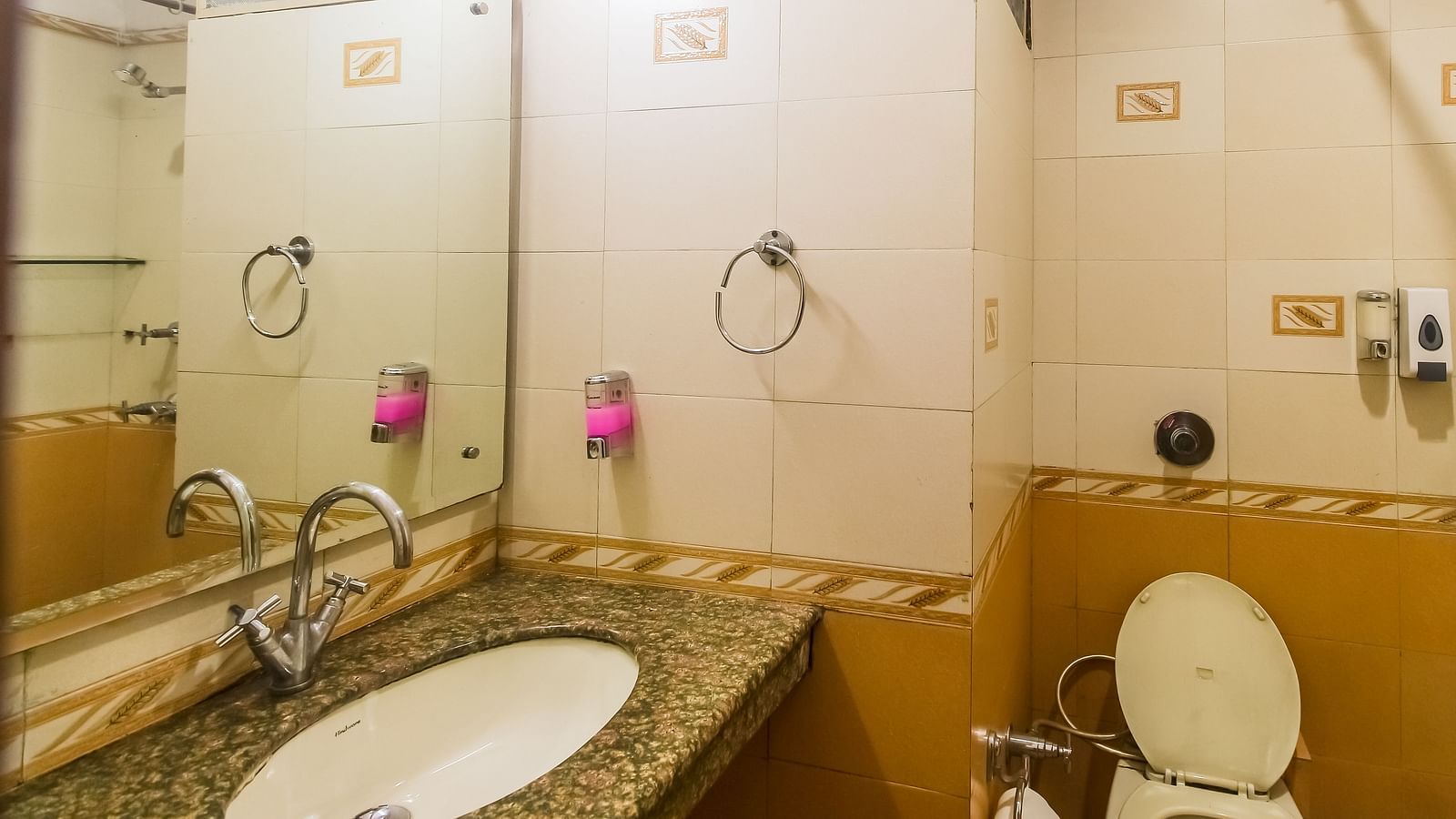 A bathroom with a white oval sink, a granite countertop, a toilet, and a tiled wall with a mirror, illuminated by a light fixture. - Hotel Diamond Plaza, Chandigarh.
