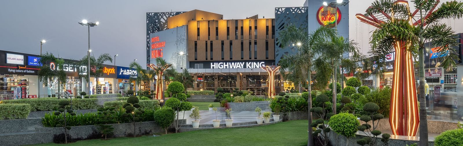 A modern building with a prominent sign is surrounded by a lush green lawn, palm trees, and outdoor lighting, captured during twilight.