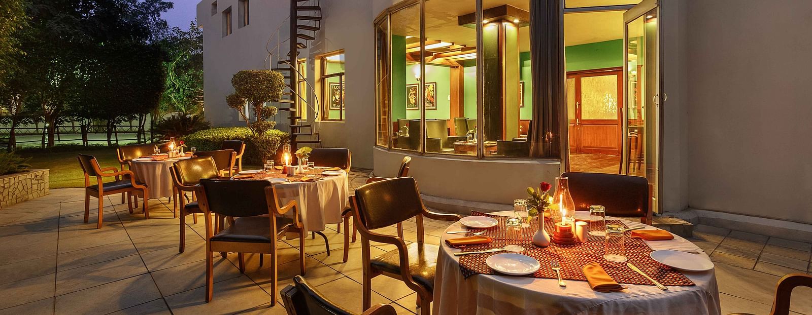 An outdoor dining setup with tables and chairs arranged outside, flanked by trees and neatly trimmed bushes