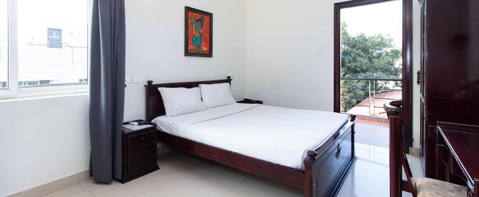 A room displaying a double bed with white bedding, a bedside table with a lamp, a wardrobe, a doorway leading to the balcony at Sanctum Suites Indiranagar, Bangalore.