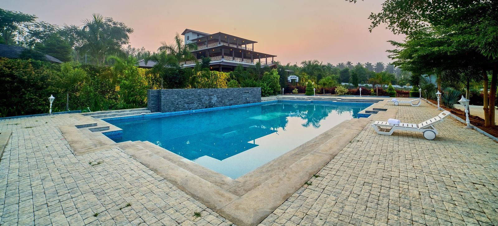 An outdoor swimming pool with a unique, geometric shape, surrounded by light-coloured paving stones and lush green trees, under a clear sky. - Vintage Trails by Ilara