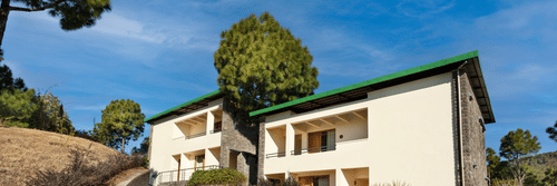 Exterior of premium rooms at Suryavilas Luxury Resort and Spa in Solan featuring the facade of the property on the mountain surrounded by trees and shrubs under a blue sky on a sunny day