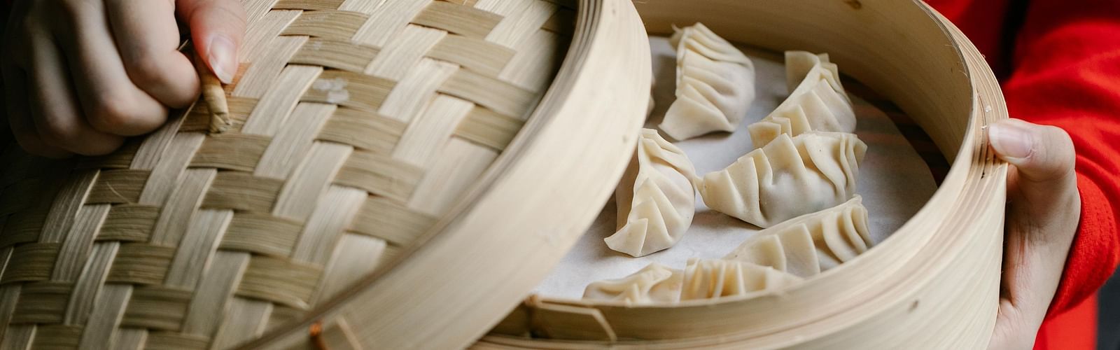 A person holding a bamboo steamer filled with dumplings, with the lid partially open